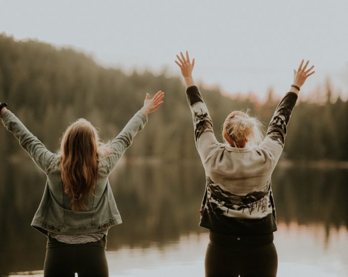 two women hands up standing beside body of water