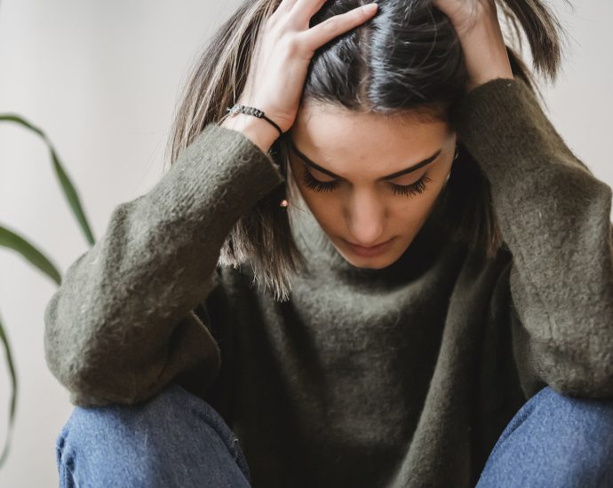 Crop disappointed young ethnic female with dark hair in casual clothes grabbing head with hands and looking down while sitting against white wall at home