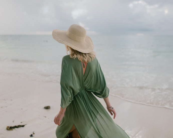 Back view of unrecognizable female tourist wearing straw hat and summer dress standing on seashore in cloudy day