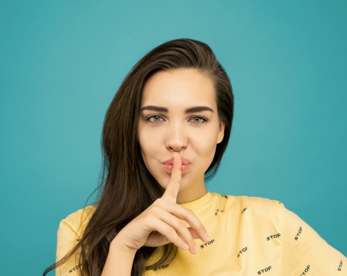 Portrait Photo of Woman in Yellow T-shirt Doing the Shh Sign While Standing In Front of Blue Background