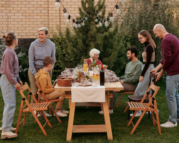 Happy family members talking and sitting down to eat tasty food at big wooden table in backyard in daytime