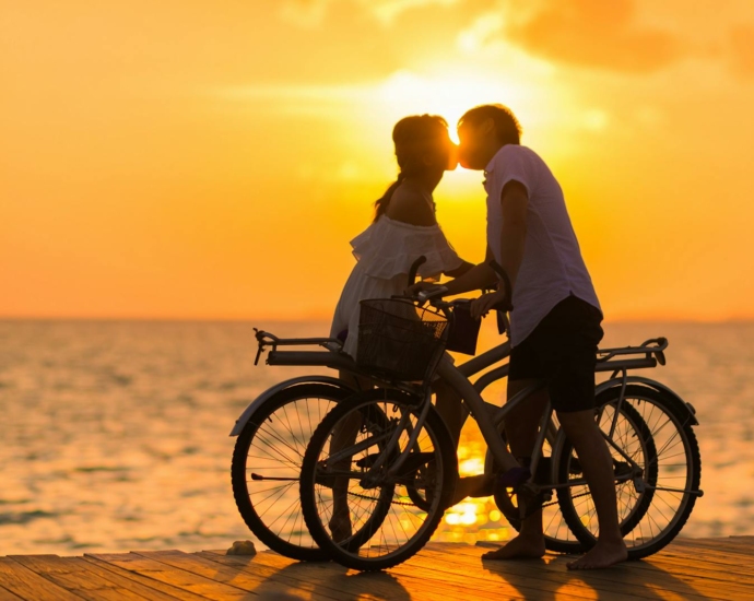 Photography of Man Wearing White T-shirt Kissing a Woman While Holding Bicycle on River Dock during Sunset