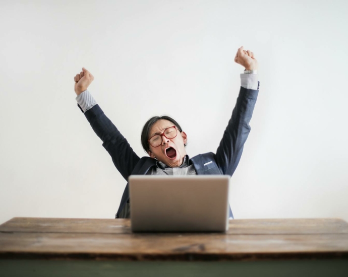 Photo of Yawning Man with His Hands Up and Eyes Closed Sitting at a Table with His Laptop