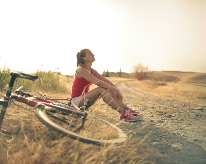 Full body of female in shorts and top sitting on roadside in rural field with bicycle near and enjoying fresh air with eyes closed