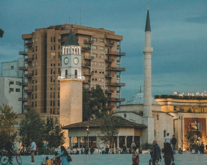 a group of people walking around a plaza with a clock tower in the background