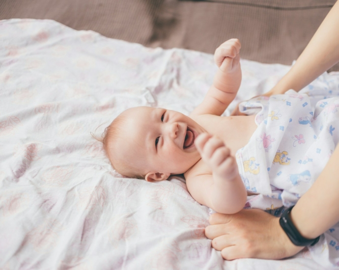 a smiling baby laying on a bed with a woman's arm