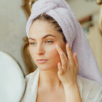 Woman with head towel applying moisturizer in front of mirror indoors.