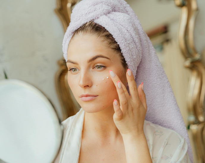 Woman with head towel applying moisturizer in front of mirror indoors.