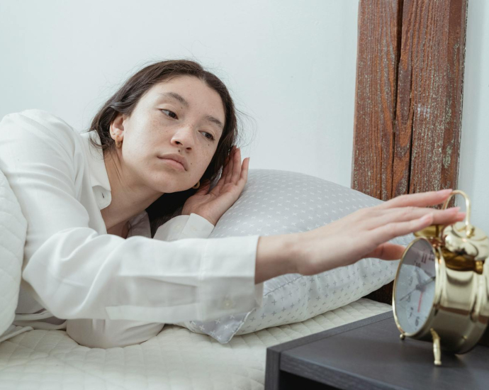 Sleepy young brunette female in white sleepwear resting in comfortable bed and waking up with alarm clock at home in morning time