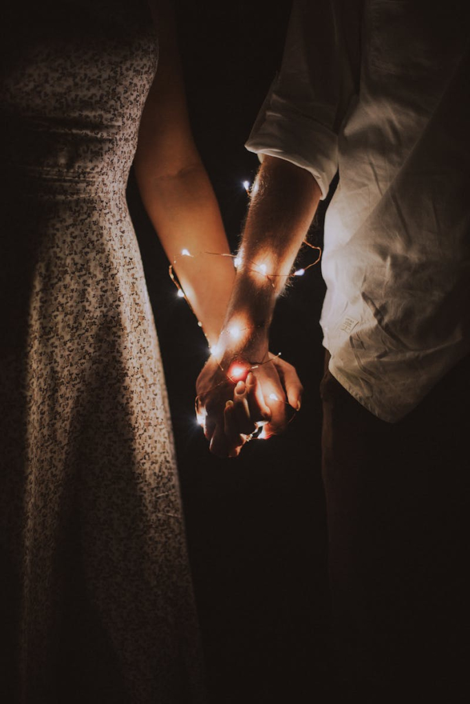 A warm and intimate close-up of a couple holding hands adorned with glowing string lights.